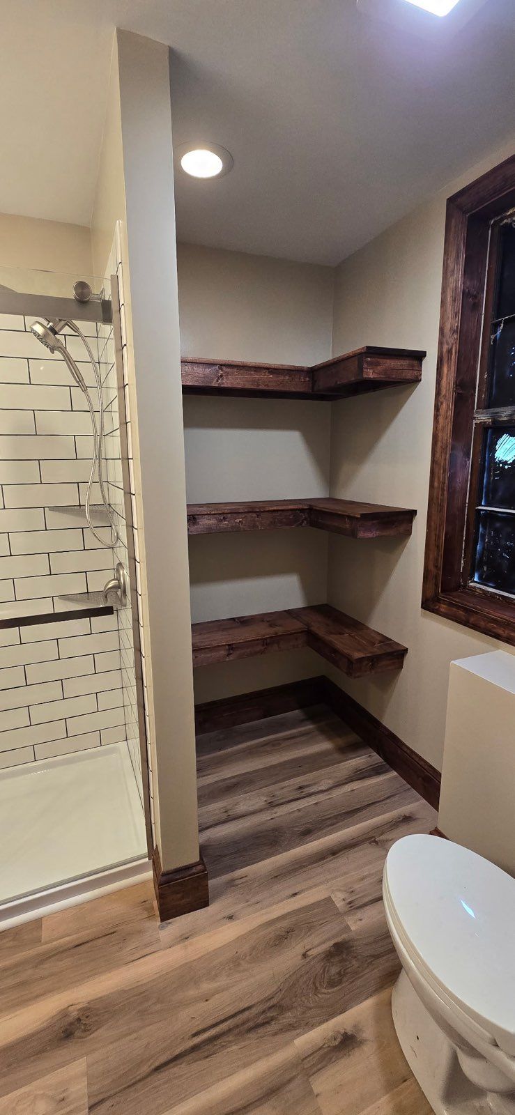 Small bathroom with corner wooden shelves, a window, toilet, and wood-look flooring.