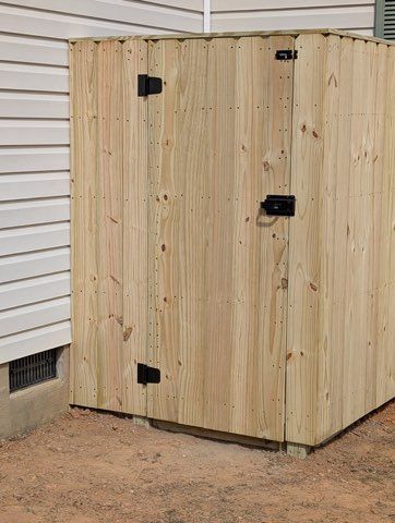 Wooden outdoor storage shed with black hinges beside a house wall on dirt ground