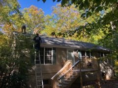 People on a ladder and roof of a brown house surrounded by leafy trees