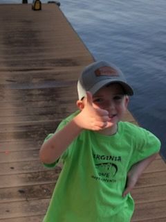 Boy in a gray cap and green shirt giving a thumbs-up on a wooden dock at sunset.