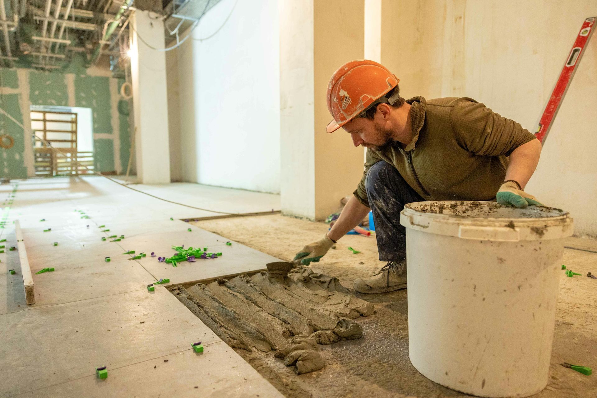 Construction worker tiling a floor, applying mortar with a trowel, wearing a hard hat, in a room under construction.