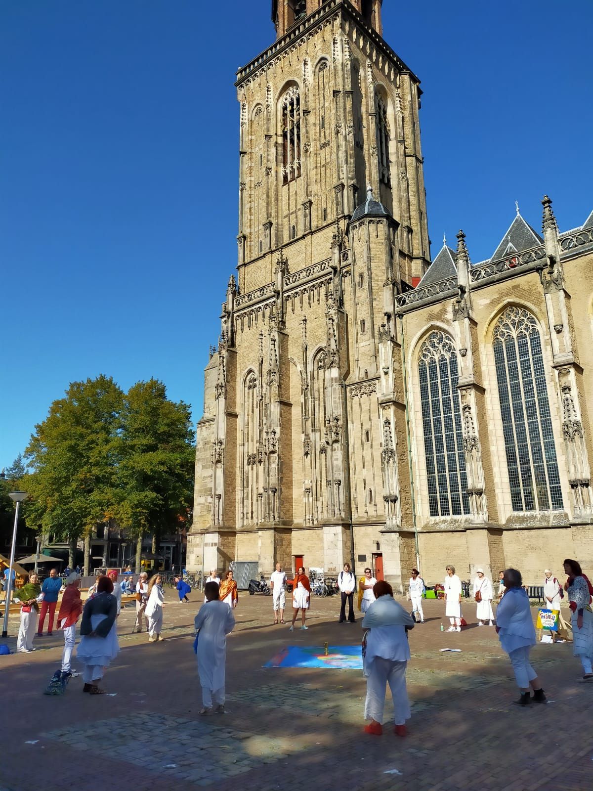 Mensen in witte kleding staan in een kring op een bakstenen plein voor een hoge, historische stenen kerk onder een blauwe hemel.