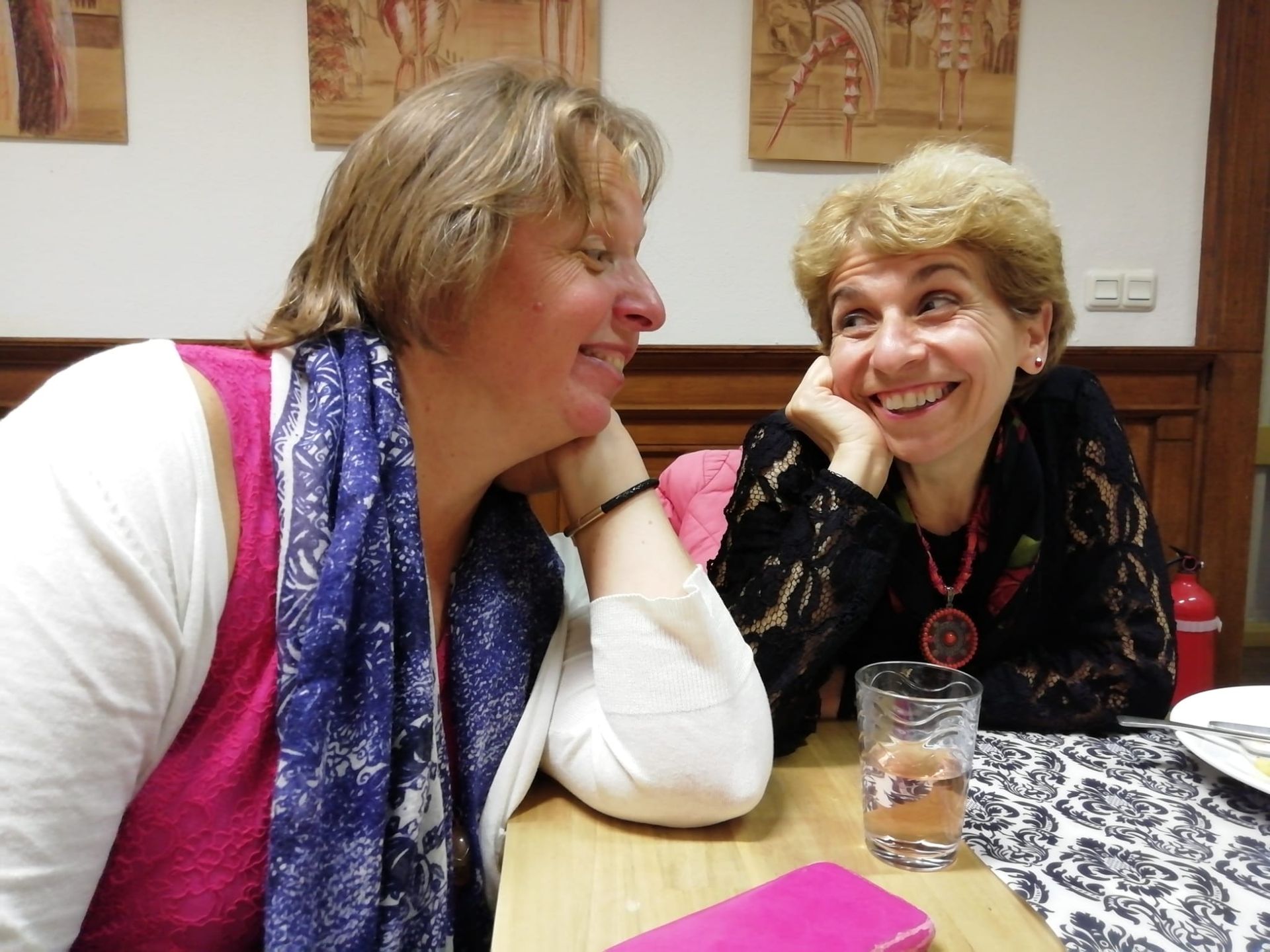 Twee vrouwen zitten aan een tafel, kijken elkaar aan en glimlachen, in een kamer met wandbekleding in diverse patronen.