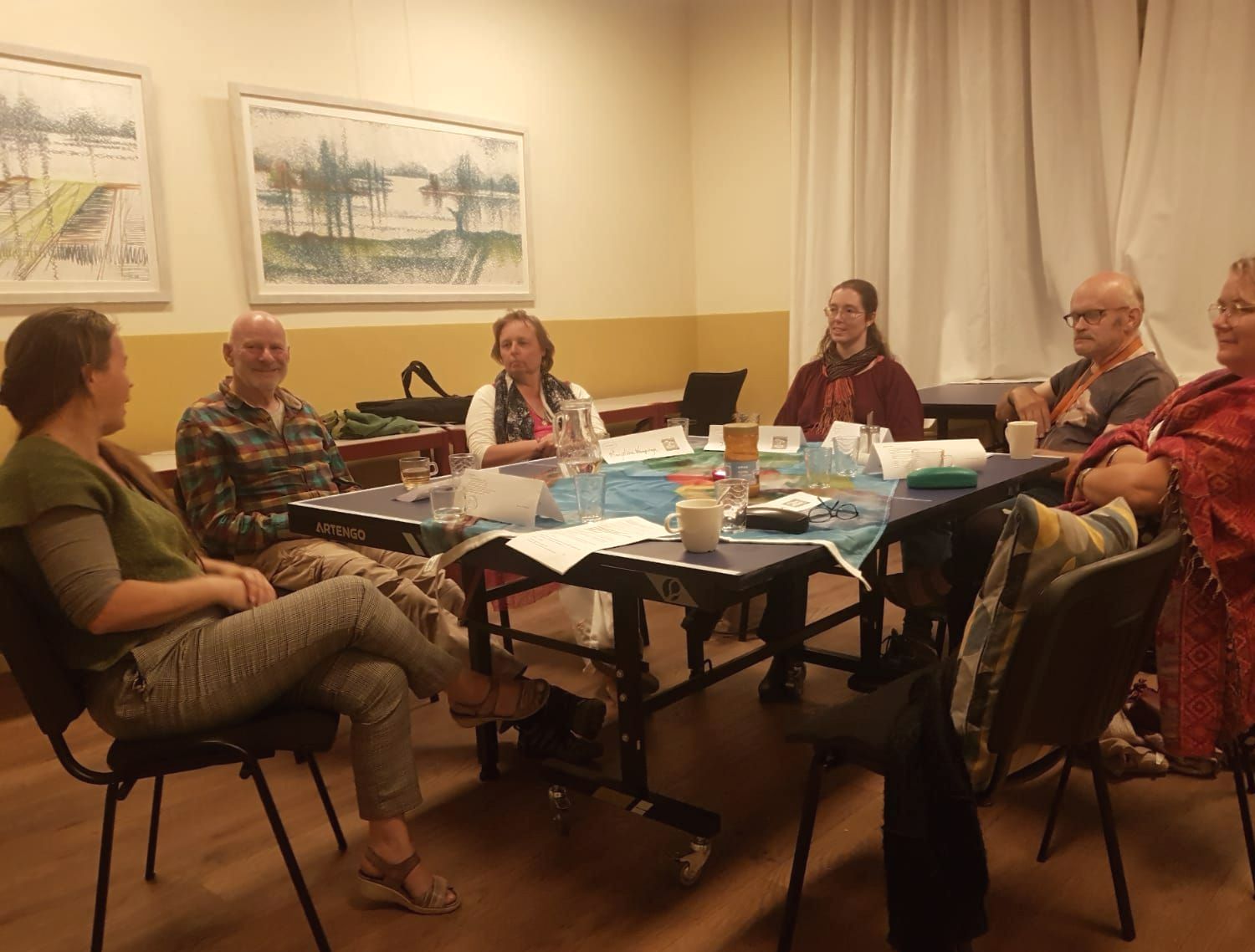 Zes mensen zitten rond een tafel in een helder verlichte ruimte voor een vergadering en voeren een ontspannen gesprek.