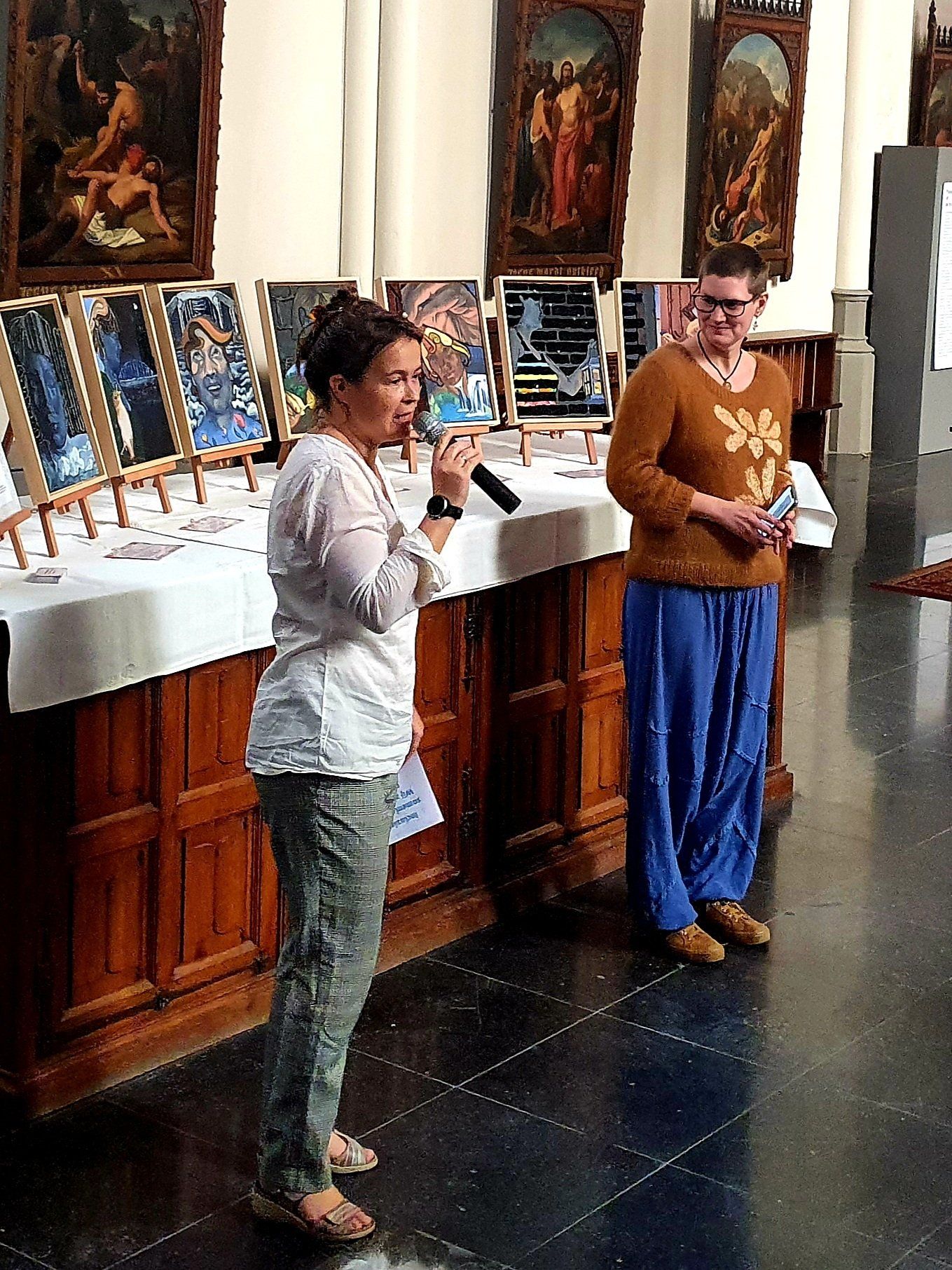 Iemand spreekt in een microfoon voor een lange tafel met tentoongestelde kunstwerken, terwijl een andere persoon er vlakbij staat.