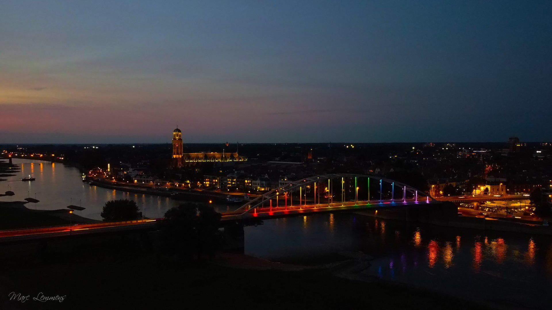 Een verlichte brug, stralend in regenboogkleuren, overspant een donkere rivier in de schemering, met in de verte de skyline van een stad.