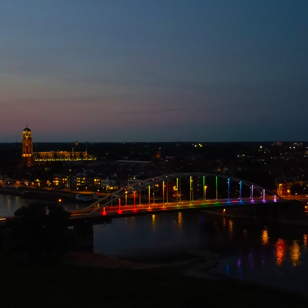 Een fel verlichte, veelkleurige boogbrug overspant een rivier in de schemering, met een kerktoren zichtbaar op de achtergrond.