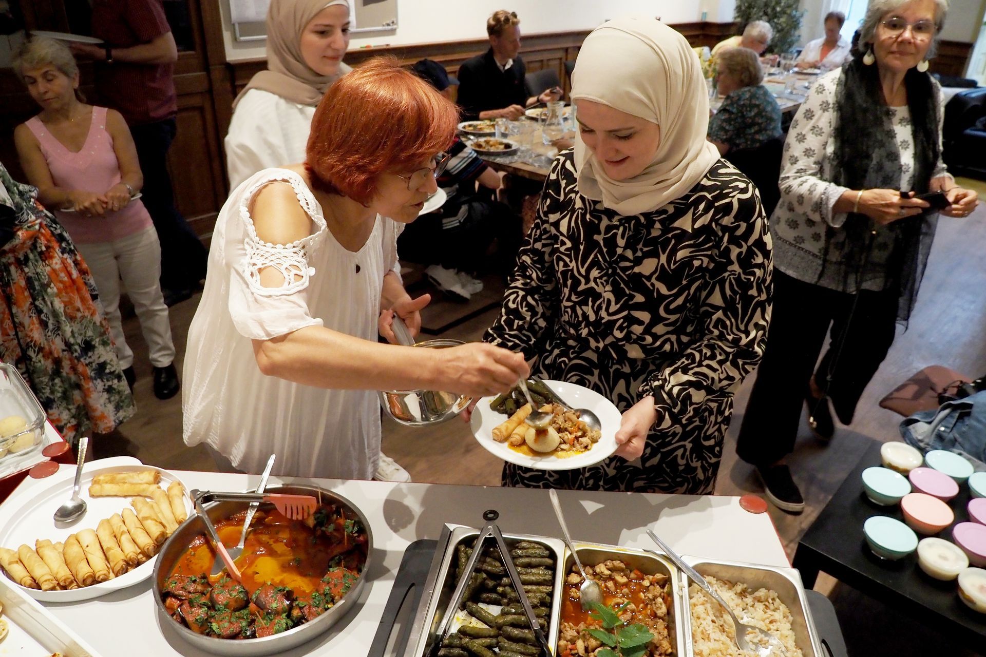 Een vrouw in een witte top serveert eten van een buffet aan een persoon in een jurk met een patroon tijdens een gemeenschappelijke bijeenkomst.