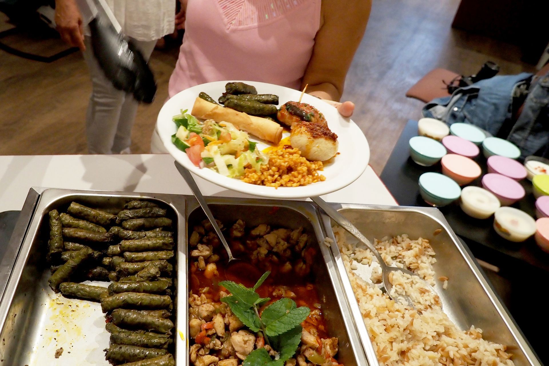 Iemand houdt een bord met eten boven een buffet met dolmades, een vleesgerecht en rijst, naast kleine kleurrijke kopjes.