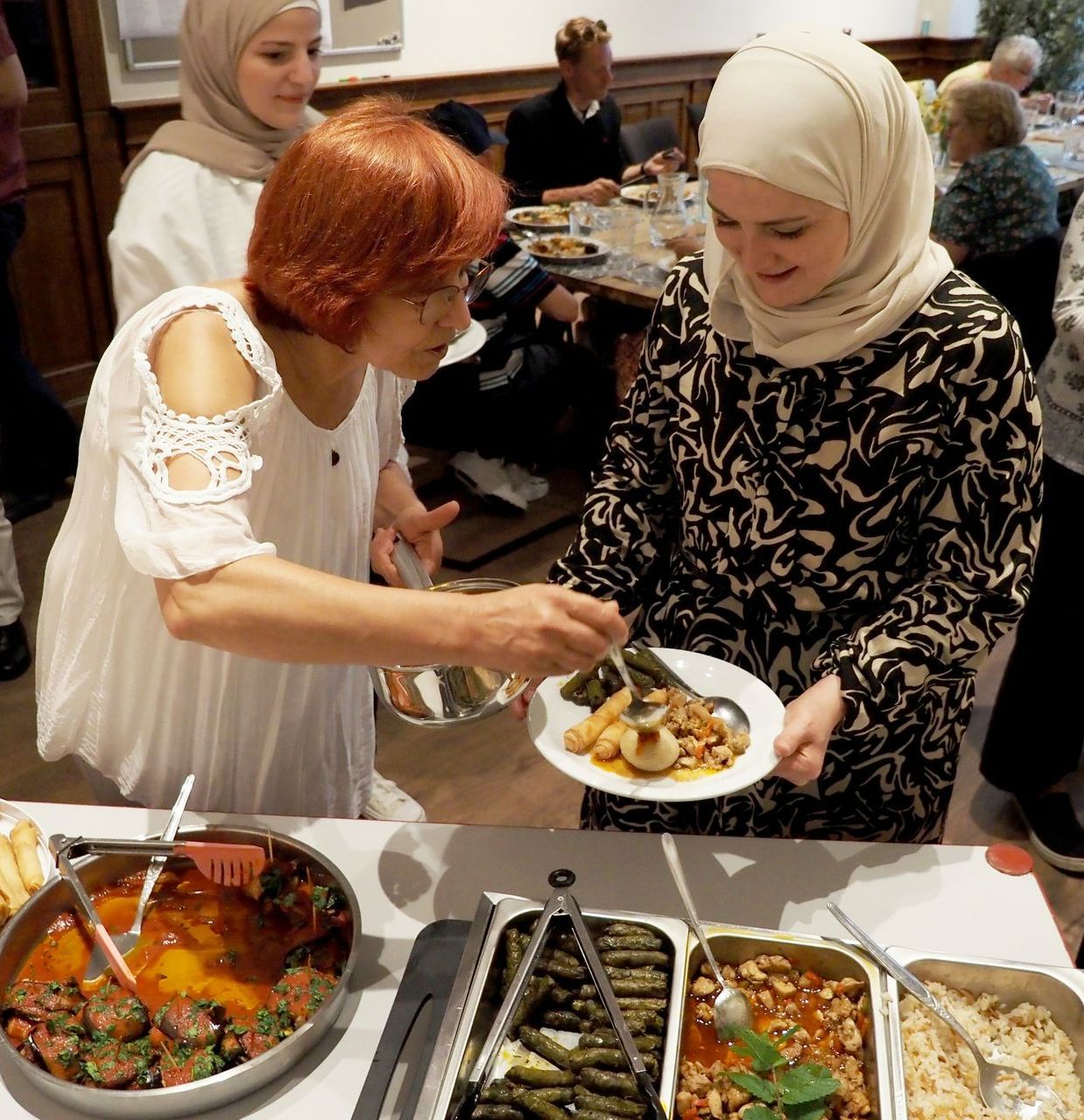 Twee mensen serveren eten vanaf een buffettafel tijdens een bijeenkomst; schalen met klaargemaakte gerechten zijn zichtbaar op het tafelkleed.