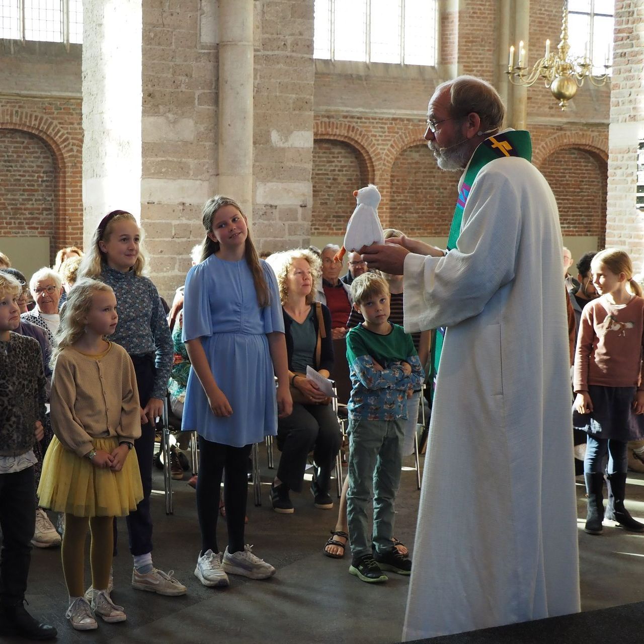 Een priester in een wit gewaad houdt een klein wit kartonnen figuurtje vast terwijl hij een groep kinderen toespreekt in een stenen kerk.