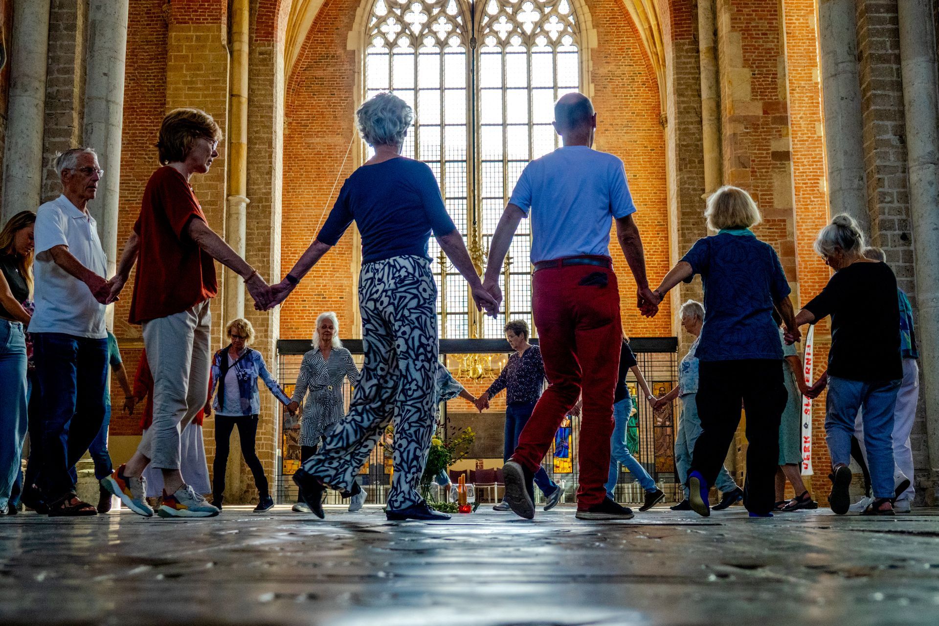 Een groep mensen, hand in hand, danst in een kring in een historisch stenen gebouw met een groot raam.