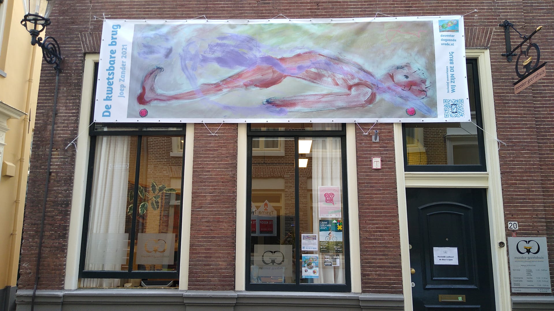 Een bakstenen winkelpui met een groot horizontaal spandoek waarop een abstracte figuur in zachtroze en paarse tinten is afgebeeld.