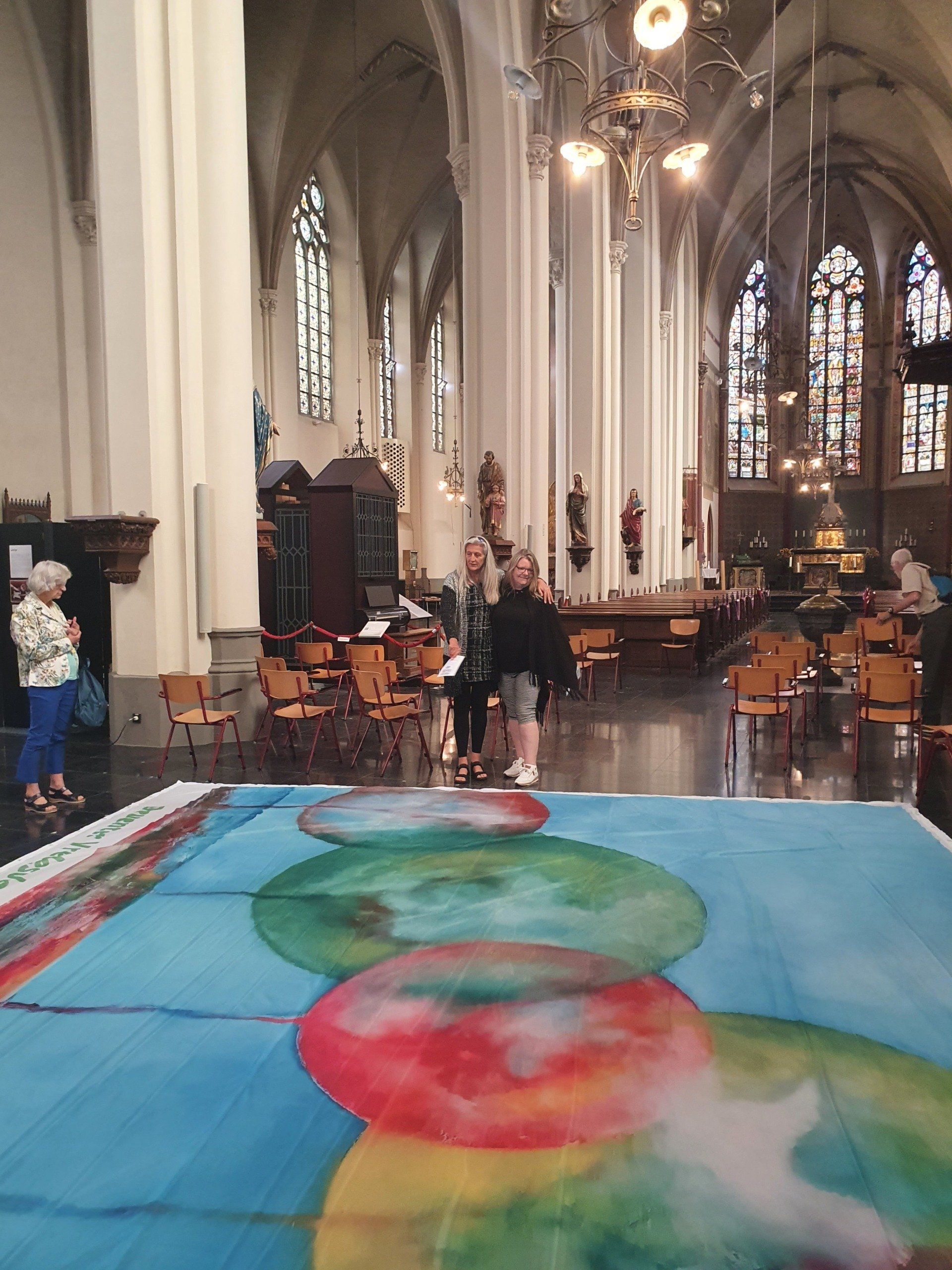 Een groot, kleurrijk abstract schilderij op de vloer van een kerk, met mensen die er vlakbij op de achtergrond staan.