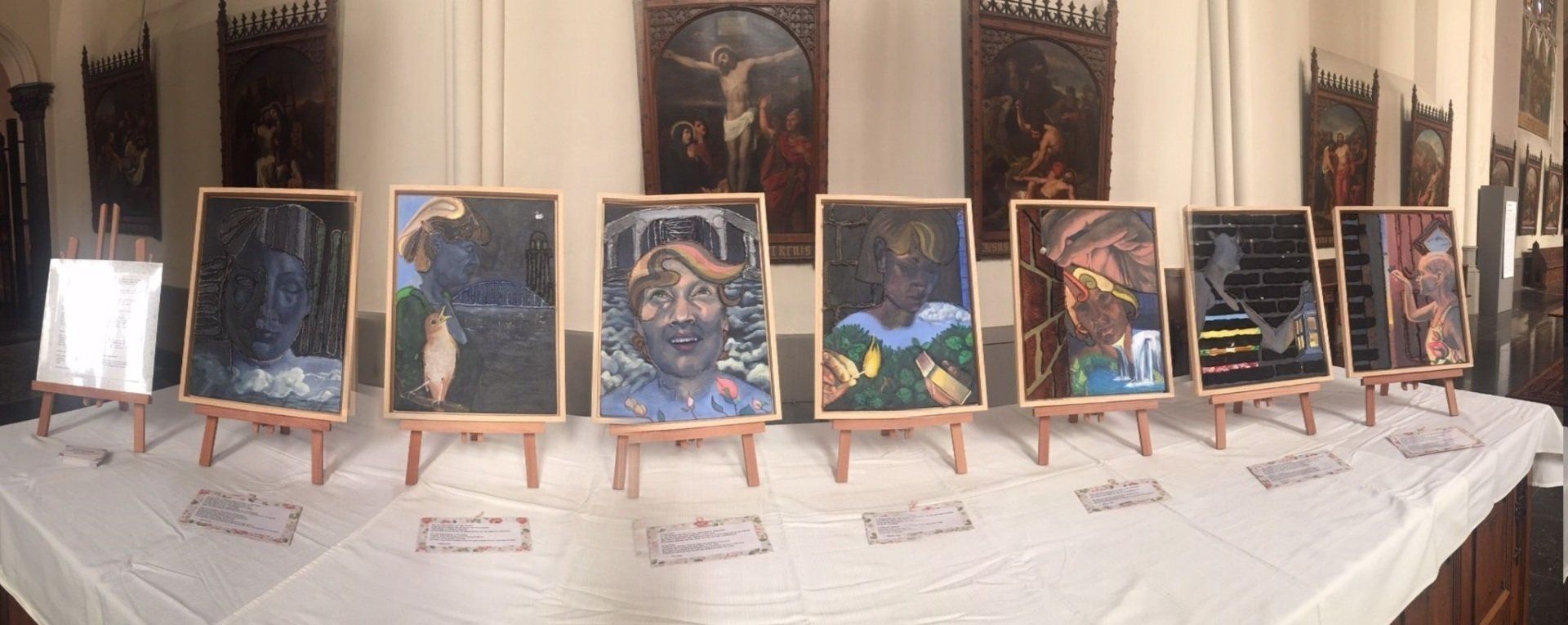 Een rij ingelijste schilderijen op kleine schildersezels, opgesteld op een met een wit tafelkleed bedekte tafel in een kamer die is versierd met religieuze kunstwerken.