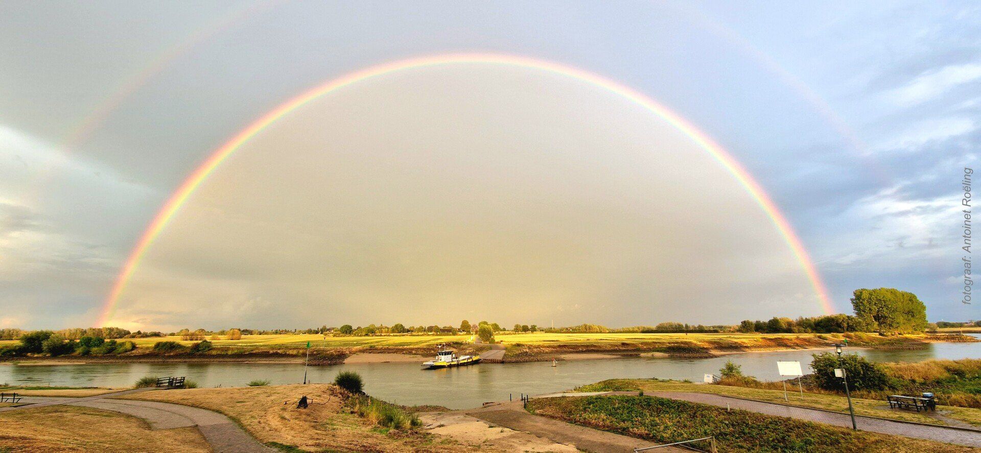 Een schitterende dubbele regenboog strekt zich uit over de IJssel bij Zutphen en een grasveld onder een bewolkte hemel.