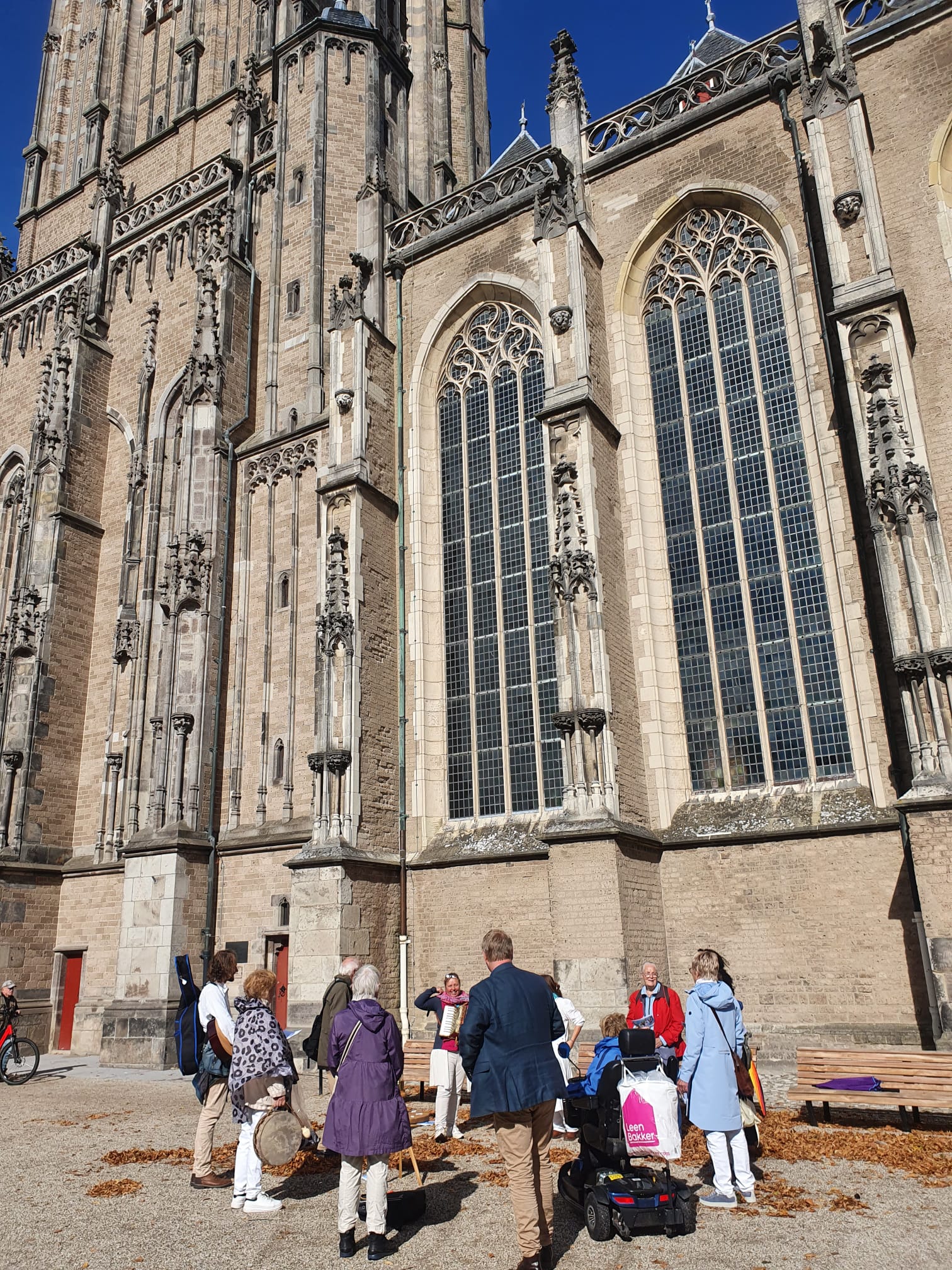 Een groep mensen verzamelt zich voor een rondleiding buiten een hoge, sierlijke gotische kerk, opgetrokken uit lichtgekleurde steen.