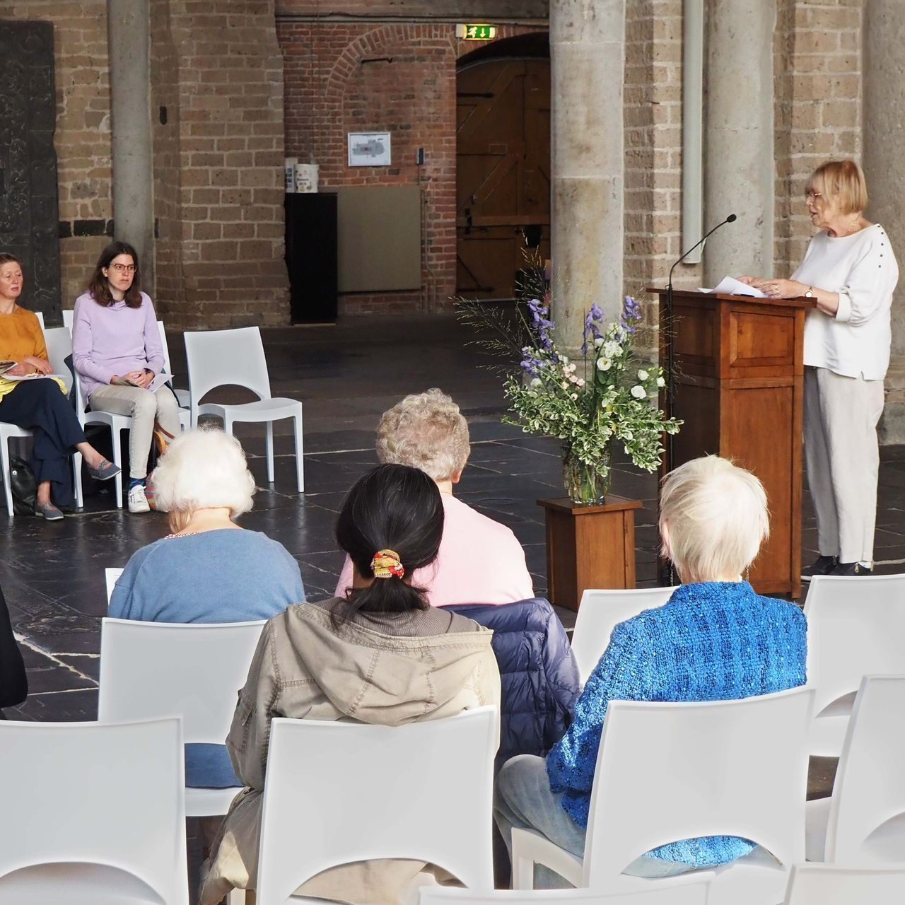 Een persoon staat op een podium en geeft een presentatie aan een zittend publiek in een stenen zaal met een bloemstuk.