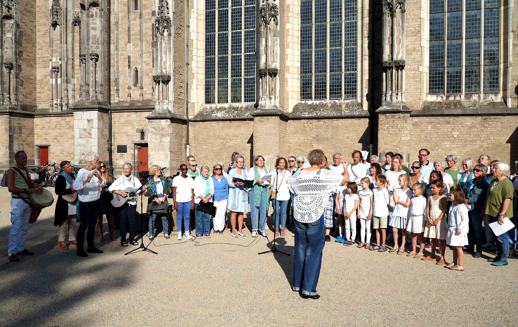 Een groep mensen zingt buiten een groot stenen kerkgebouw, met een dirigent tegenover hen en muzikanten links van hen.