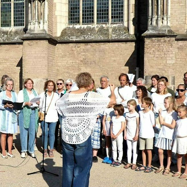 Een groep mensen staat buiten een stenen gebouw te zingen, terwijl iemand hen vanaf de voorkant dirigeert.