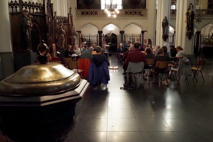 Een groep mensen zit op stoelen in een kerk, met op de voorgrond een groot, gepolijst bronzen doopvont.