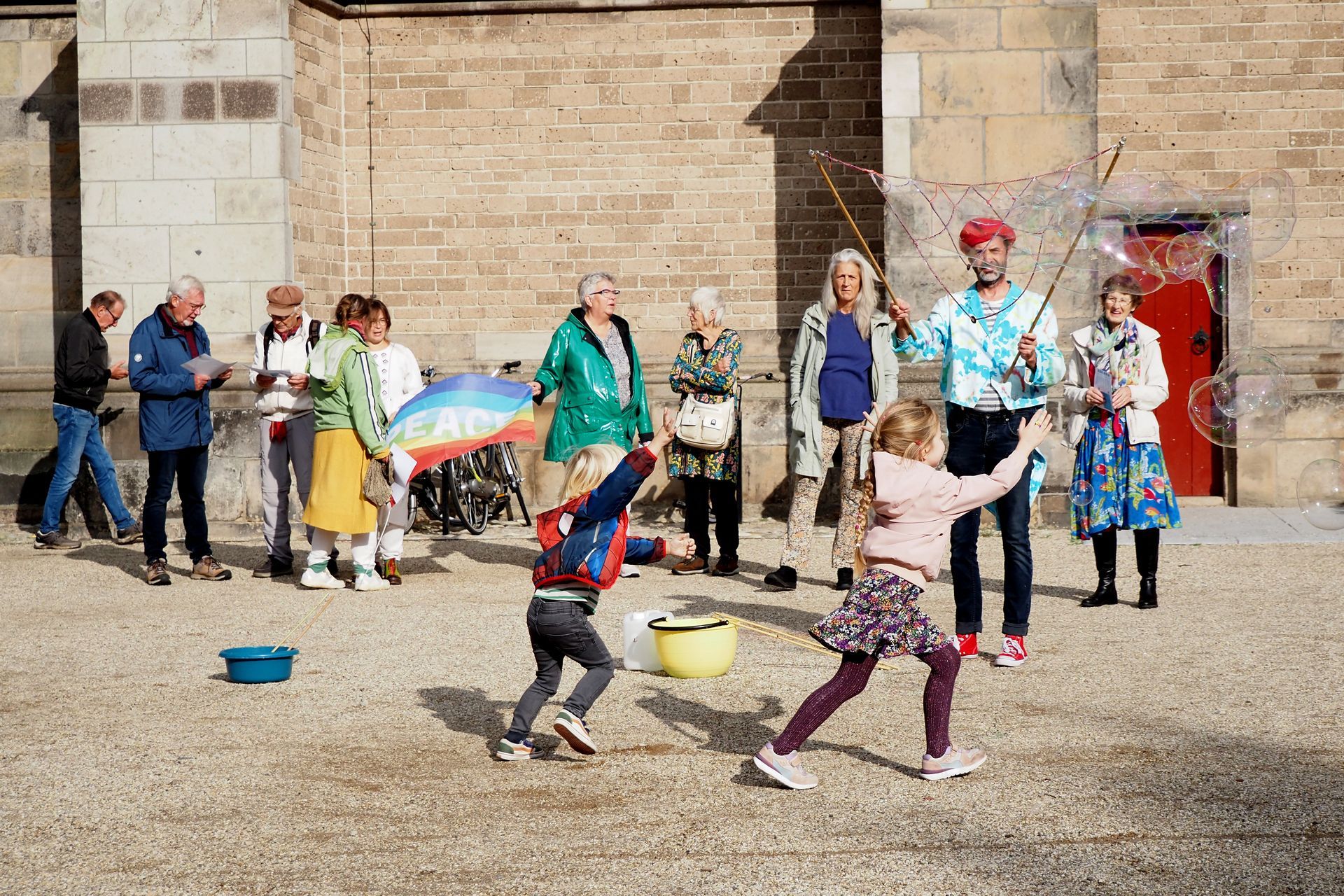 Iemand maakt gigantische bellen voor kinderen die spelen op een zonnig stenen plein voor een gebouw, onder toeziend oog van een groep.