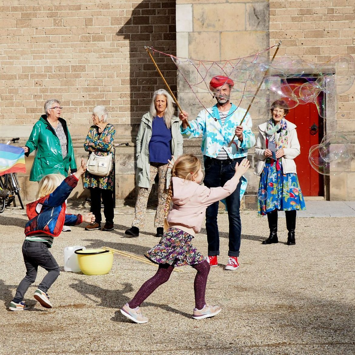 Een entertainer maakt gigantische bellen voor een stenen gebouw, terwijl een kind zijn hand uitsteekt en een groep mensen toekijkt.