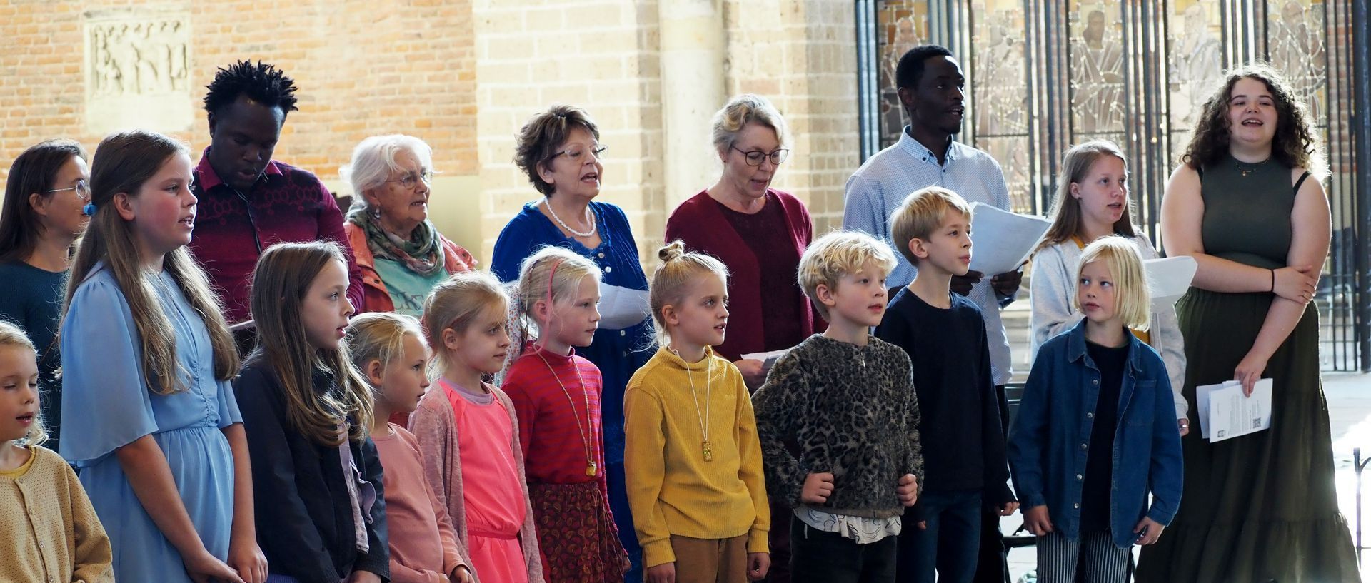 Een diverse groep zangers staat in de Lebuinuskerk en treedt op met geconcentreerde gezichtsuitdrukkingen.