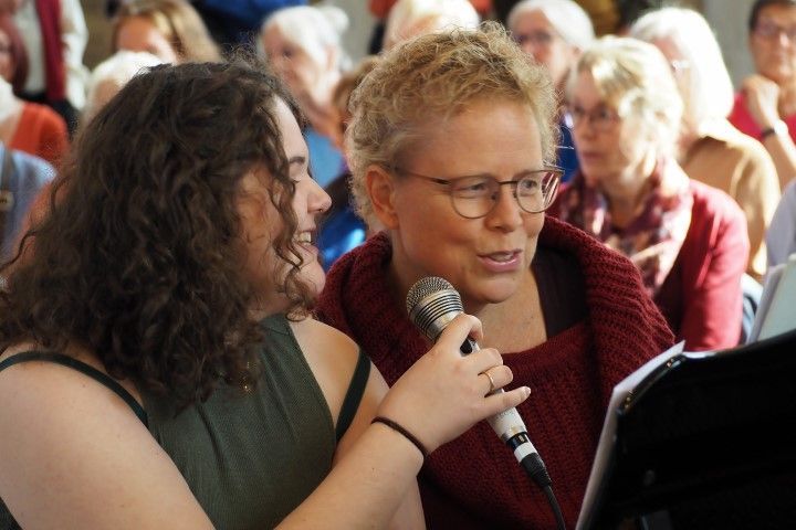 Twee mensen zingen samen in een microfoon, terwijl ze naar een bladmuziek kijken in een zaal vol zittende bezoekers.
