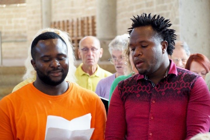 Twee personen op de voorgrond zingen terwijl ze papieren vasthouden, omringd door een groep in de Lebuinuskerk.