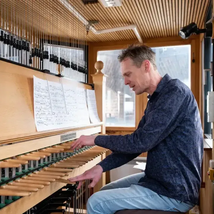 Iemand zit achter een carillonklavier en bespeelt de houten hendels met zijn handen terwijl hij de bladmuziek leest.