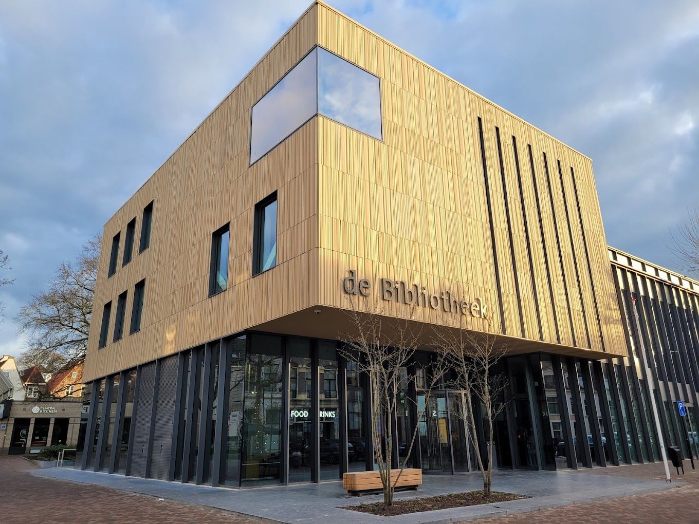 Modern bibliotheekgebouw met een gevel van licht hout met een decoratief patroon, donkere verticale kolommen op de begane grond en glazen wanden.