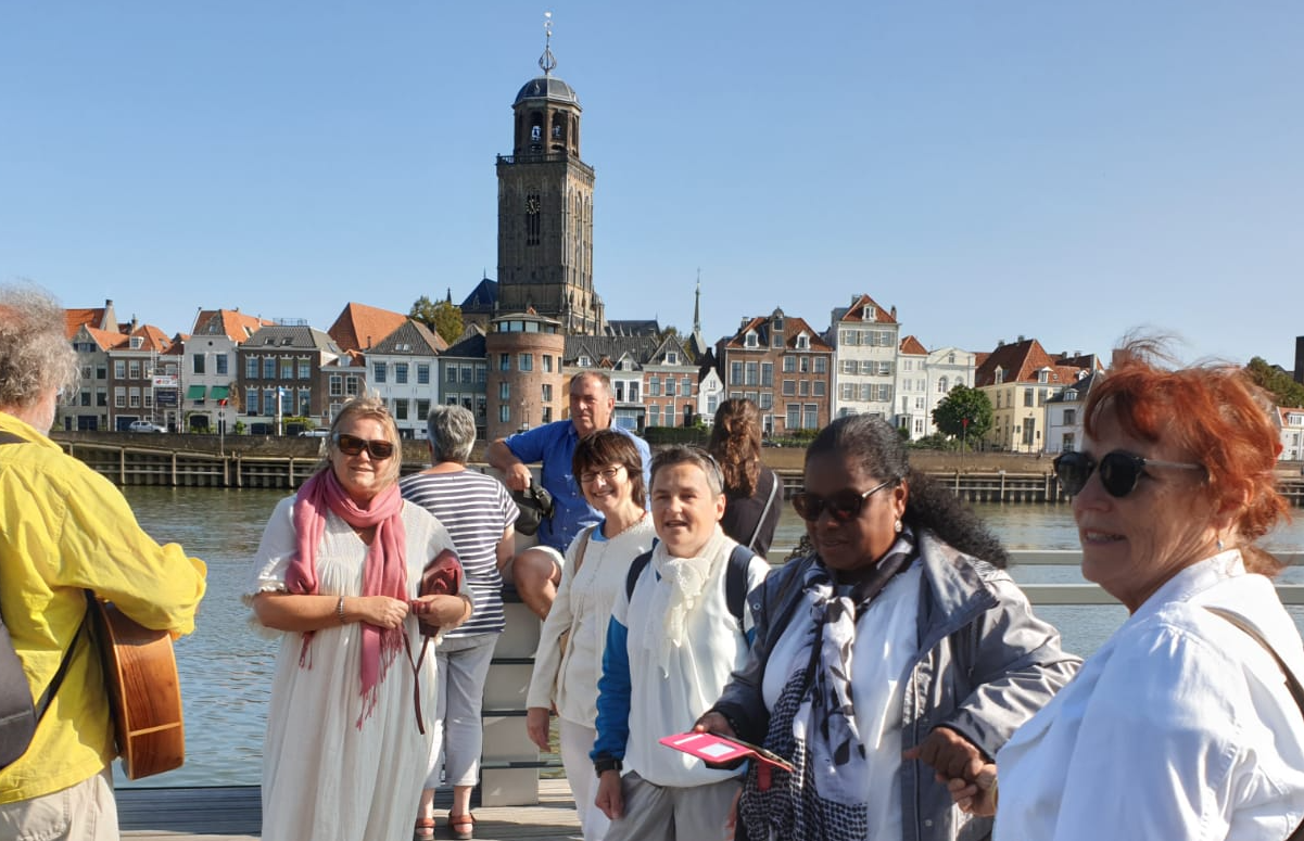 Een diverse groep toeristen staat op een pier aan het water met op de achtergrond een historische stad en een klokkentoren.