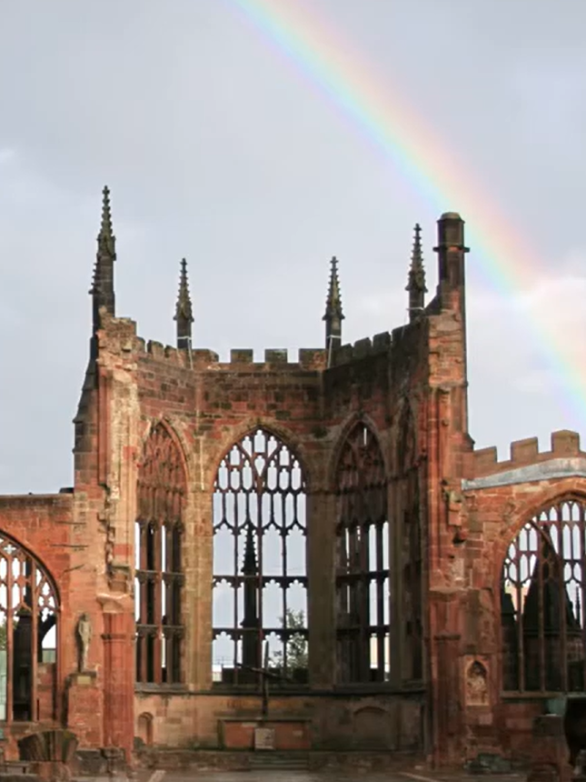 Een regenboog boog zich over de ruïnes van de zandstenen muren en lege gotische ramen van de kathedraal van Coventry tegen een bewolkte hemel.
