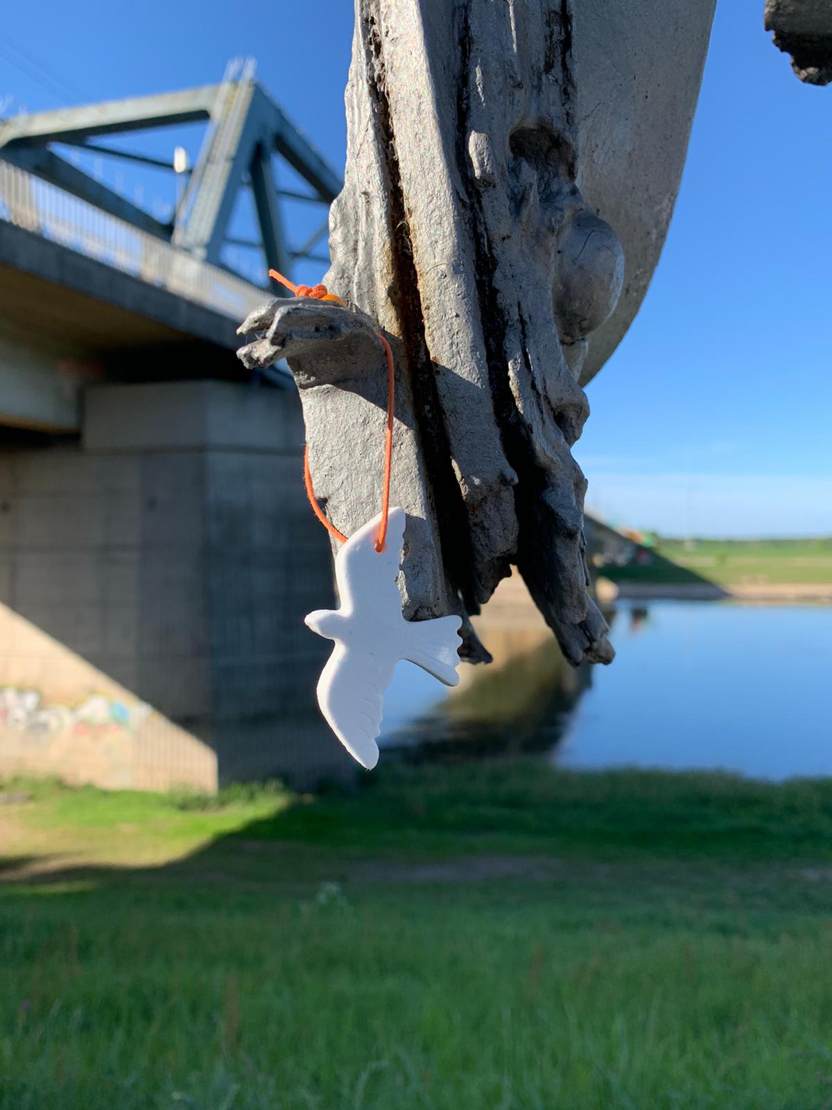 Een klein, wit keramisch vogeltje hangt aan een oranje touwtje aan een verweerde, grillige betonnen constructie vlakbij een brug.