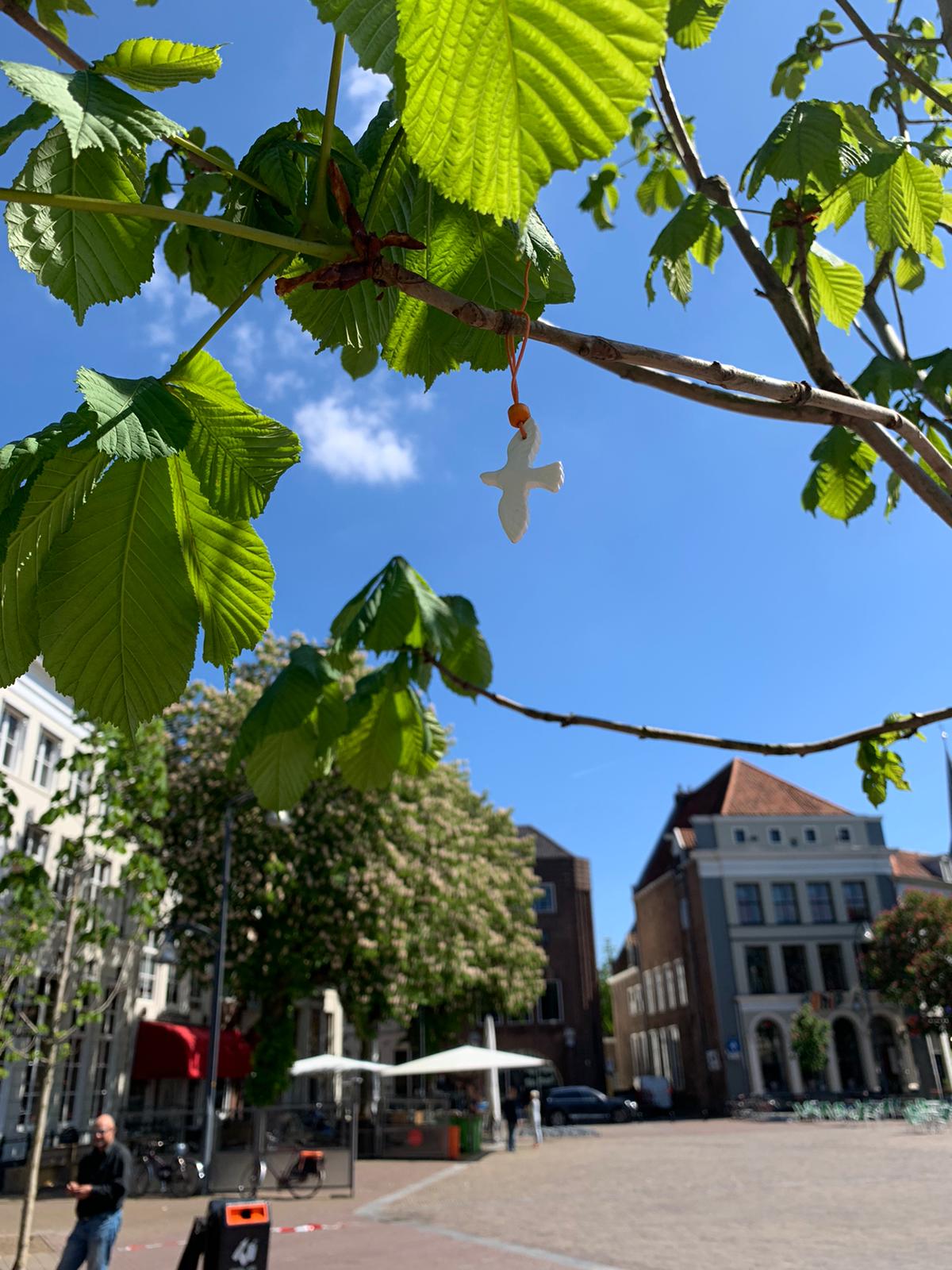 Een wit vogelornament hangt aan een bladerrijke boomtak op een zonnig stadsplein met gebouwen op de achtergrond.