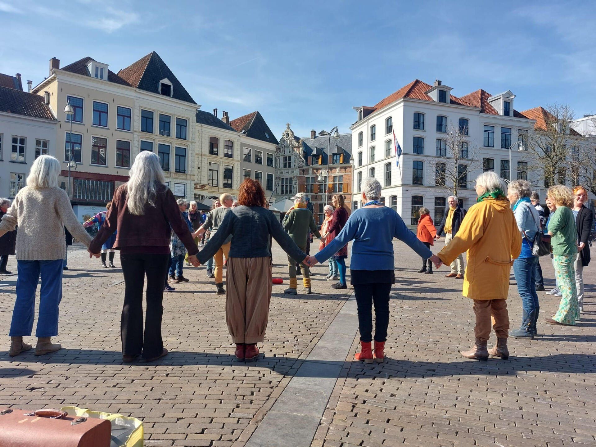 Een groep mensen staat in een grote cirkel, hand in hand, op een geplaveid plein omringd door gebouwen in Europese stijl.