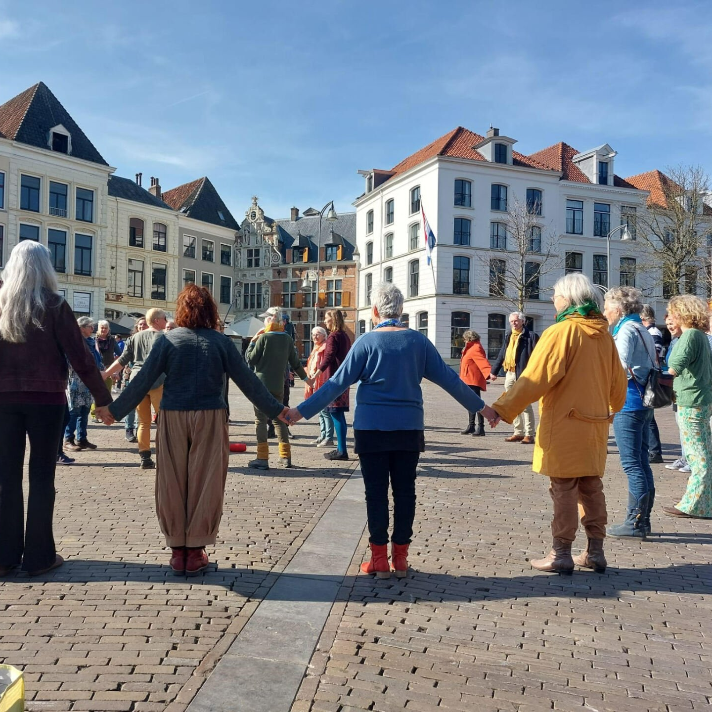 Een diverse groep mensen staat hand in hand in een kring op een geplaveid stadsplein, omringd door historische gebouwen.
