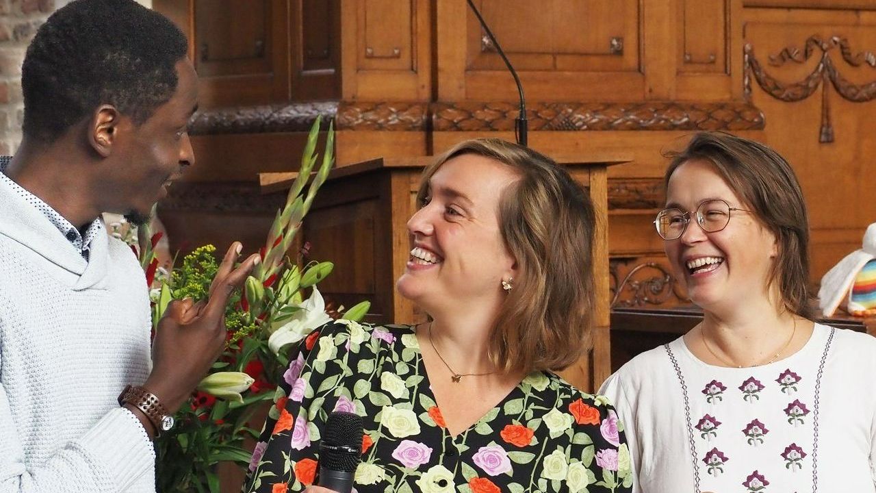 Drie mensen lachen tijdens een evenement, staand voor een houten constructie en een bloemstuk.