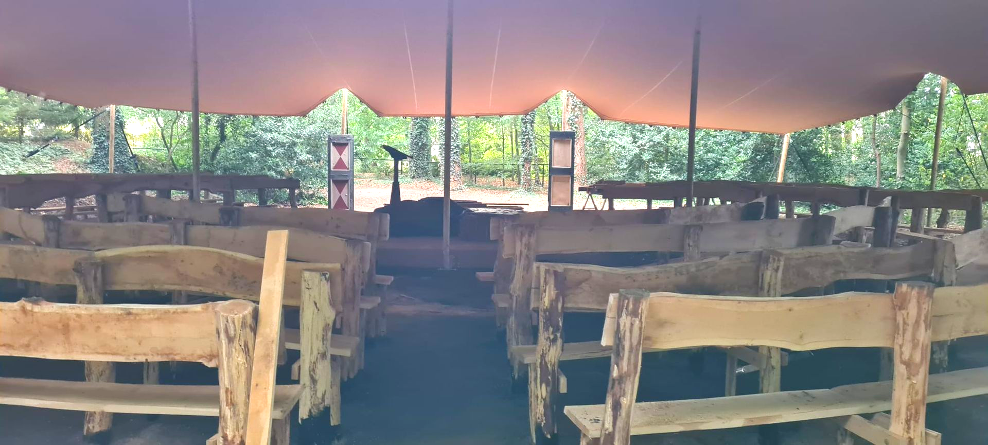 Een openluchtlocatie met rijen houten banken onder een grote beige luifel, tegenover een podium in een bosrijk gebied.