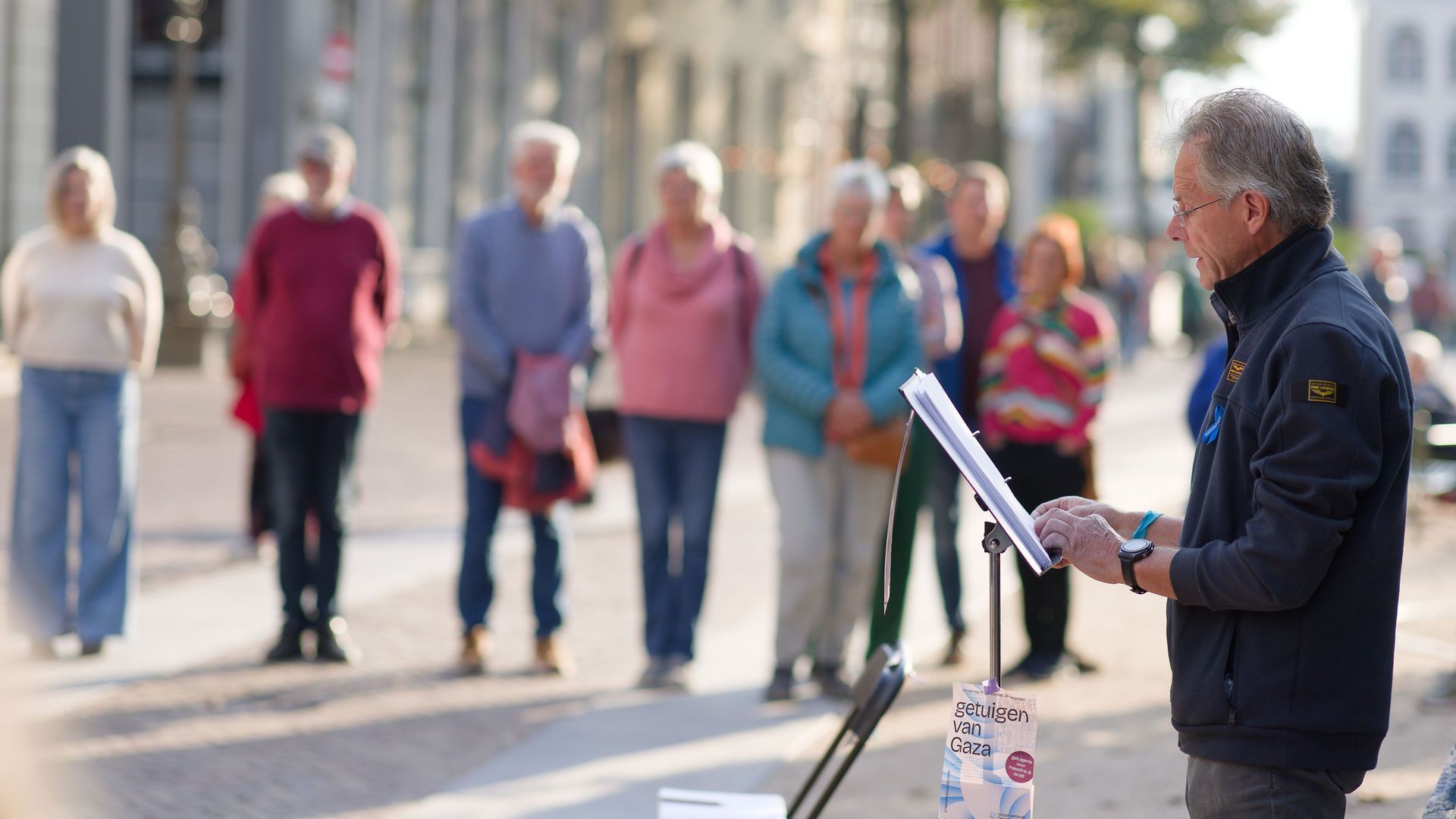 Een man in een donker vest leest voor vanaf een lessenaar aan een klein, staand publiek op een geplaveid stadsplein.