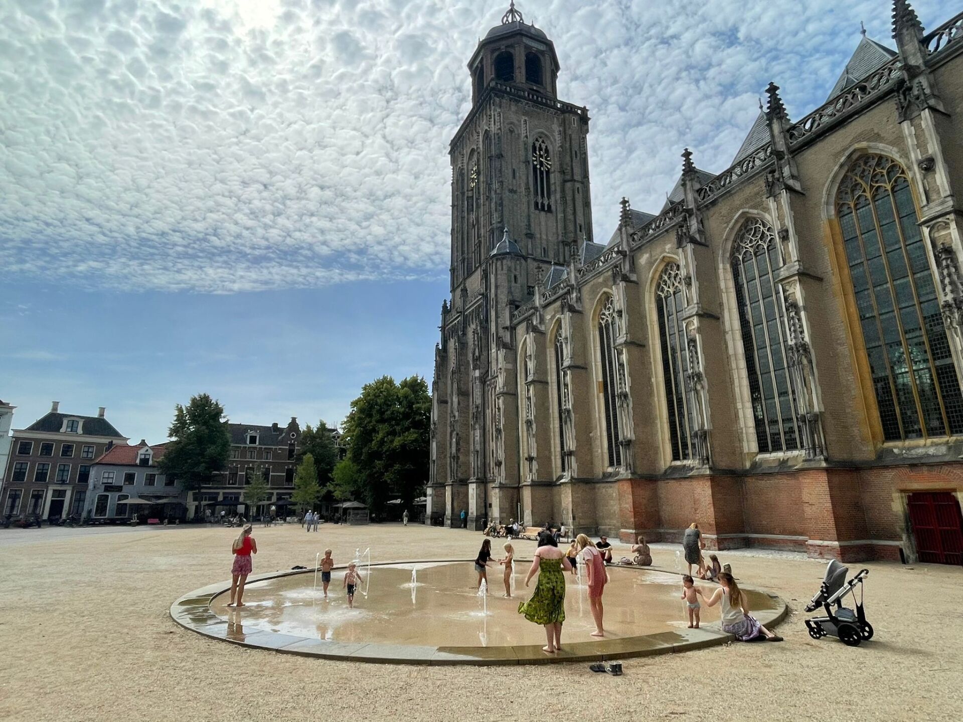 Mensen spelen in een ronde fontein op een groot, met grind bedekt stadsplein naast een hoge, historische stenen kerk, onder een bewolkte hemel.