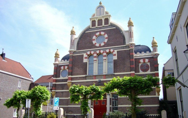 Een bakstenen synagoge met een afgeronde gevel, decoratieve bogen en rode deuren, gezien vanaf de straat tussen lommerrijke bomen.