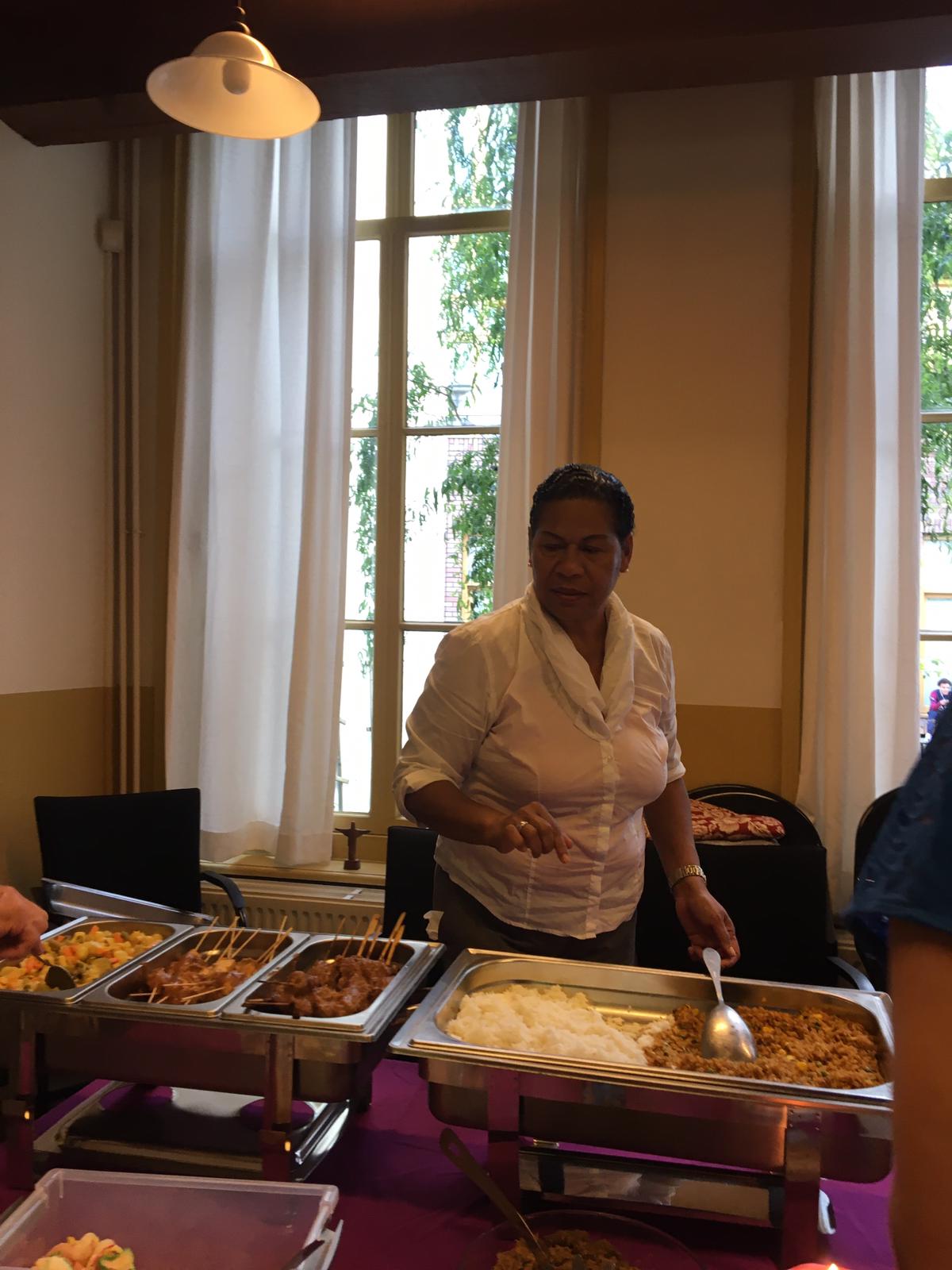Een vrouw in een witte blouse serveert eten vanaf een buffettafel met meerdere warmhoudschalen.