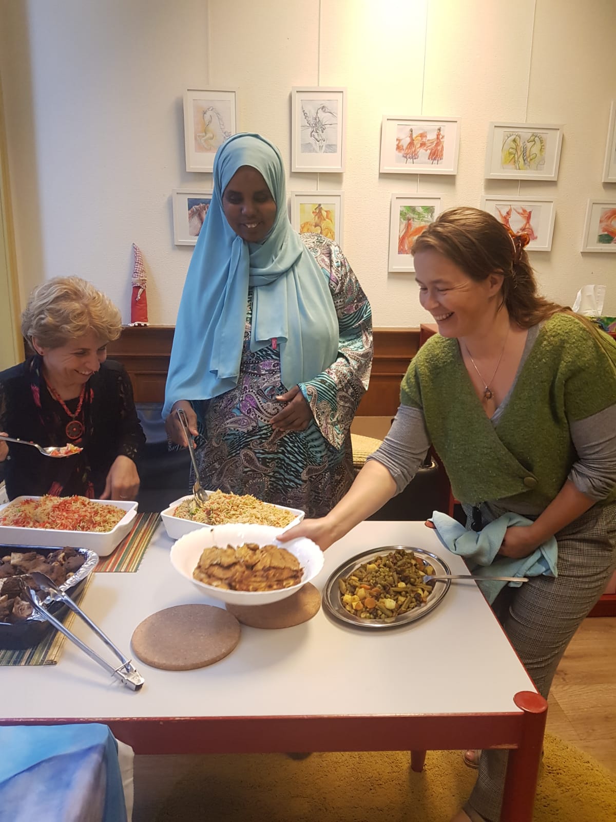 Drie mensen zitten rond een tafel vol met diverse gerechten en serveren met een glimlach het eten in een helder verlichte binnenruimte.