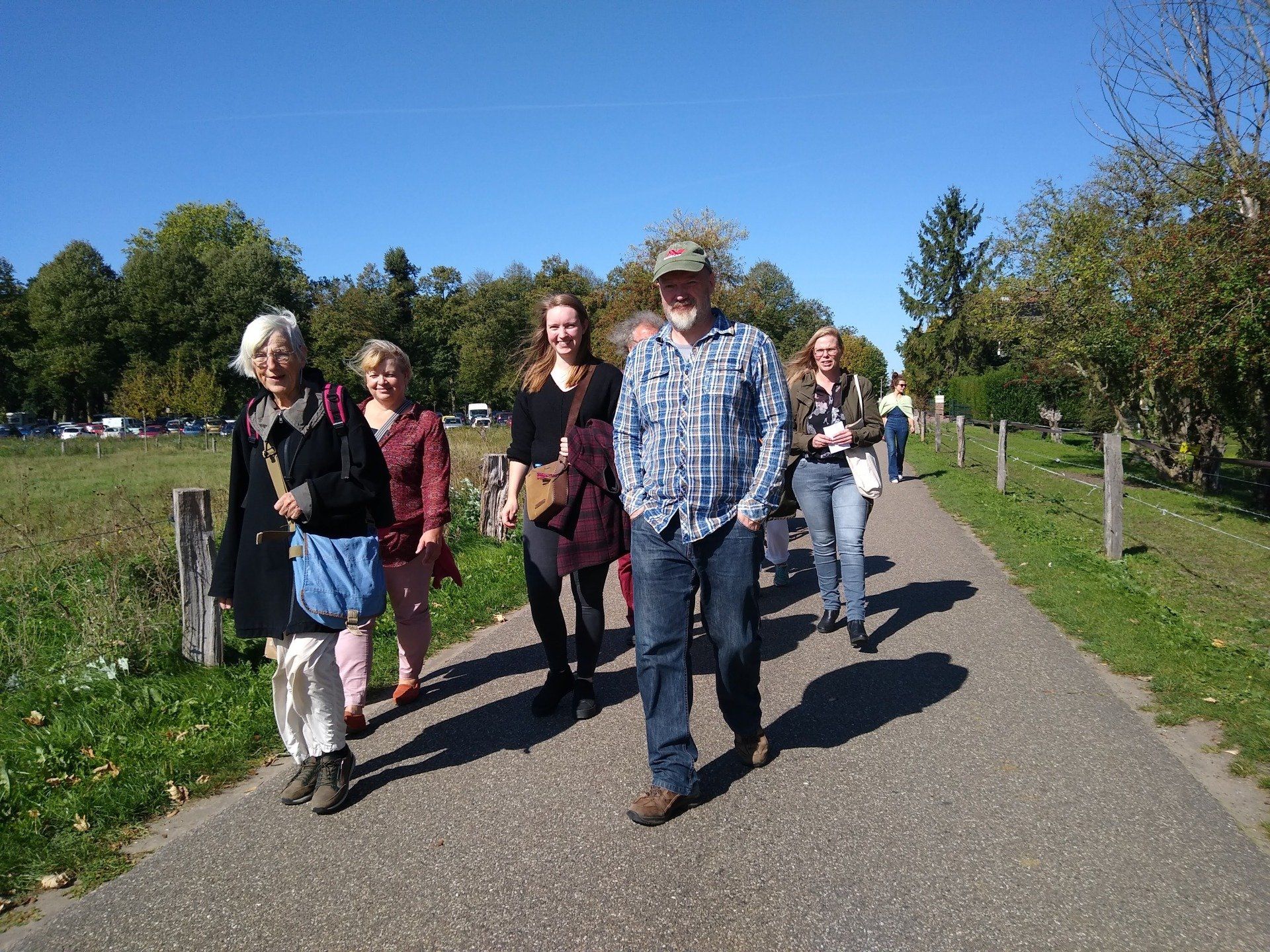 Een groep mensen loopt samen over een geplaveid pad, omzoomd door bomen en een grasveld, op een zonnige dag.