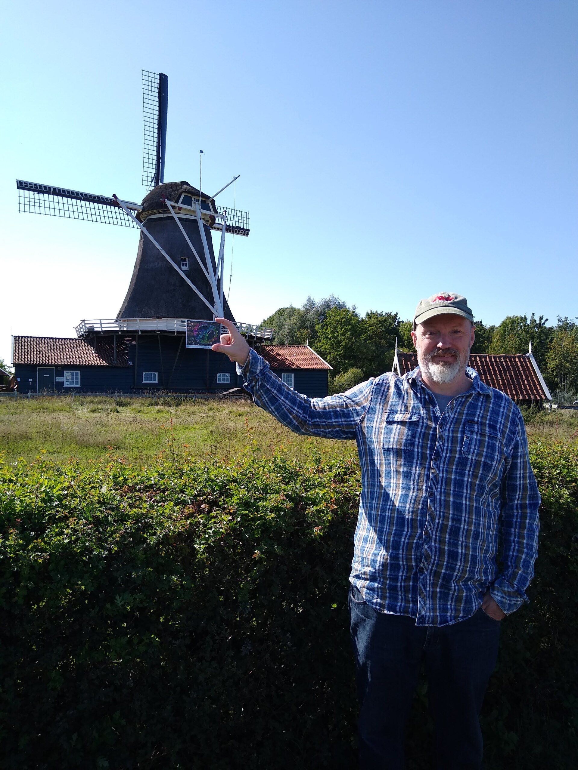 Een persoon in een blauw geruit overhemd staat voor een traditionele windmolen onder een helderblauwe hemel.