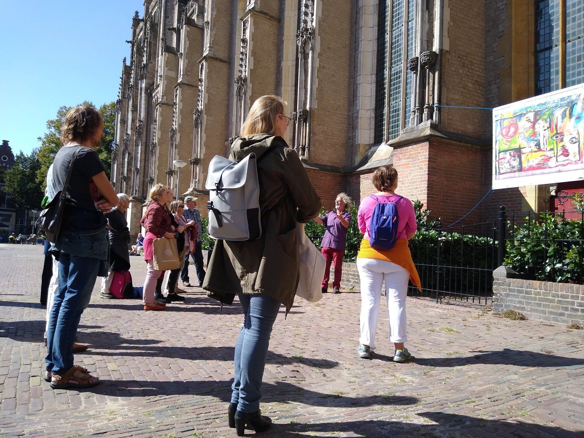 Een groep mensen staat buiten een oud bakstenen gebouw in gotische stijl en kijkt naar een poster aan de muur.