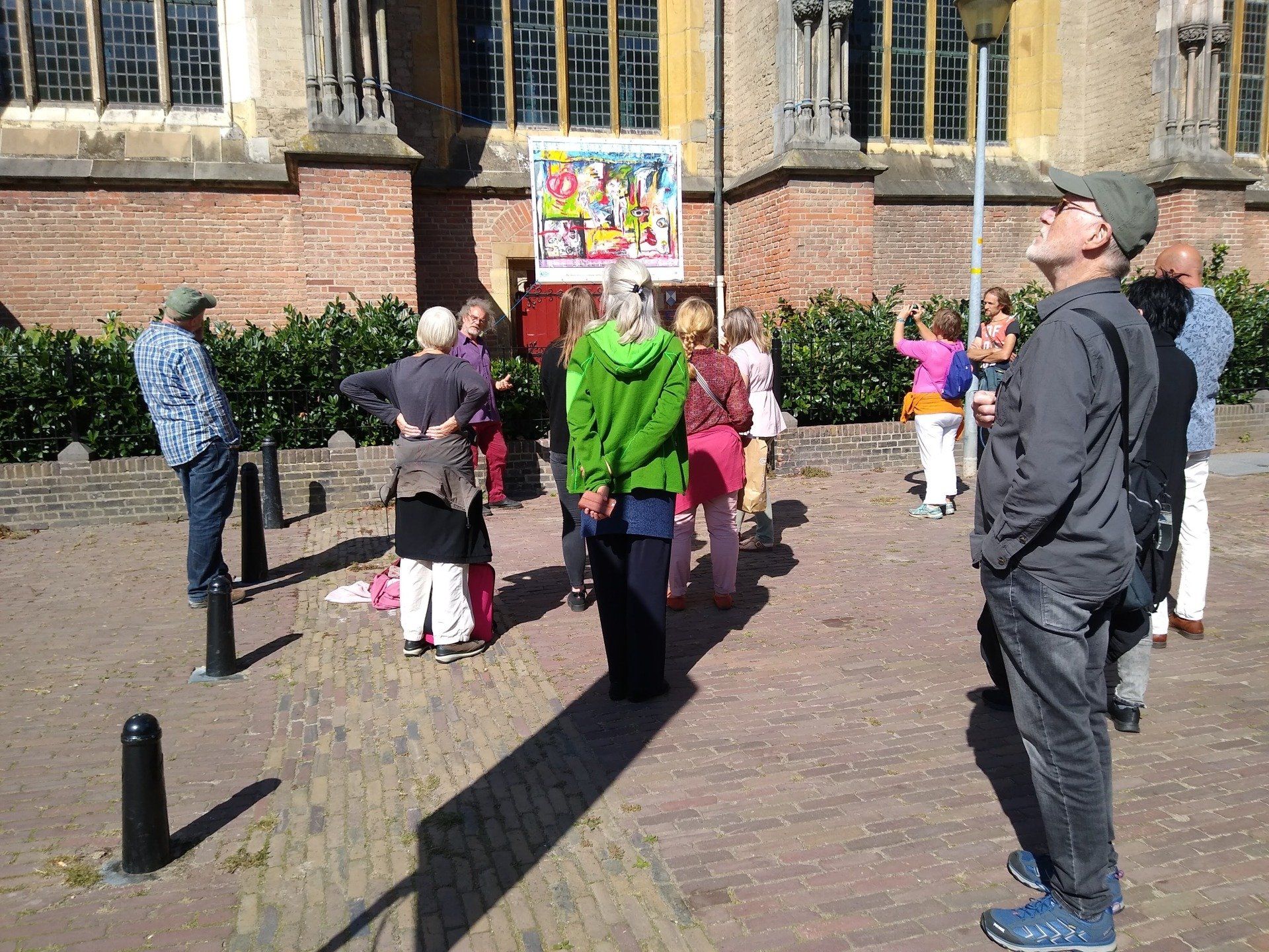 Een groep mensen staat buiten een bakstenen gebouw en kijkt omhoog naar een poster die aan de muur hangt.