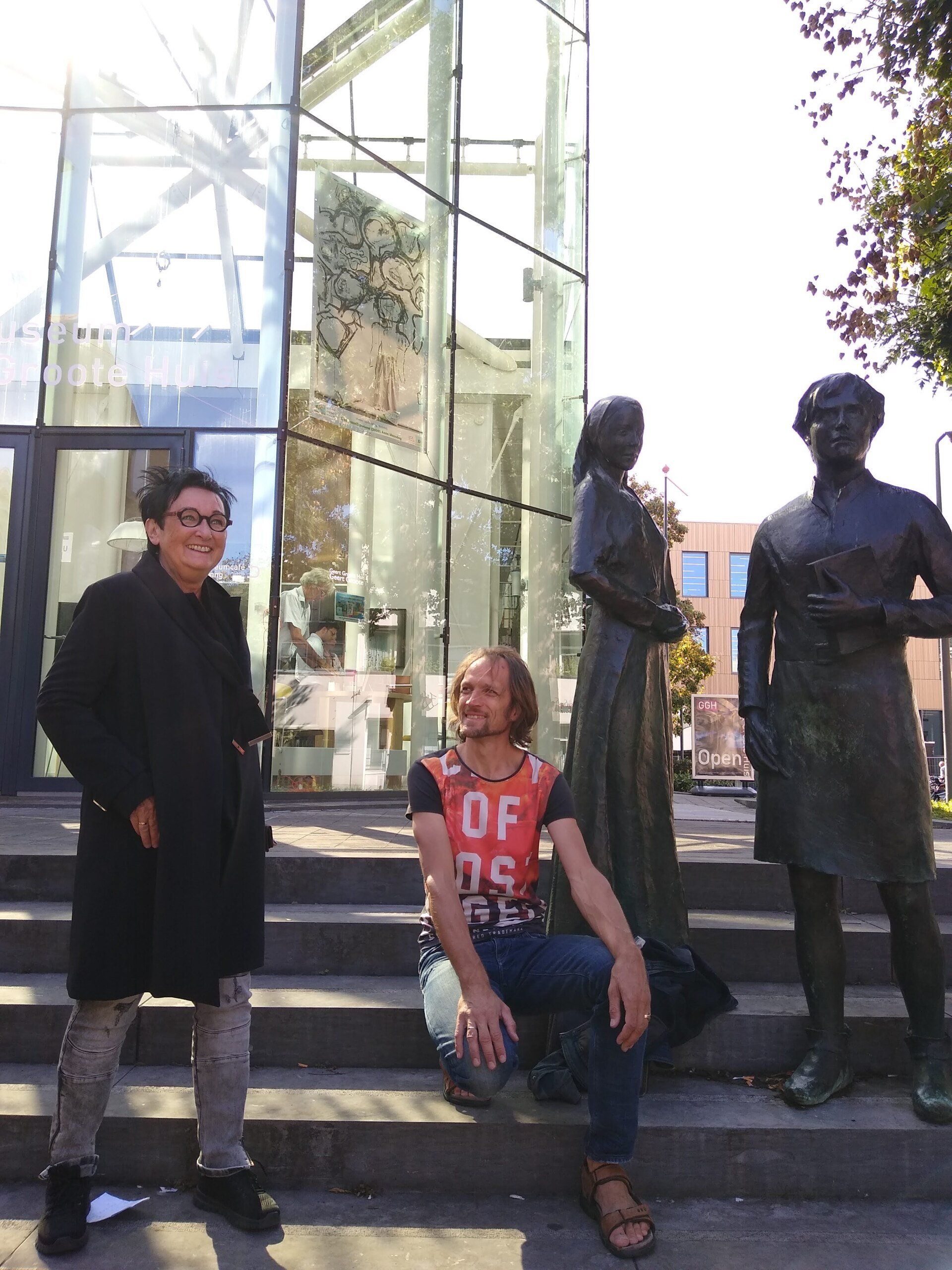 Twee mensen staan en poseren op stenen trappen voor een glazen gebouw en twee bronzen beelden.
